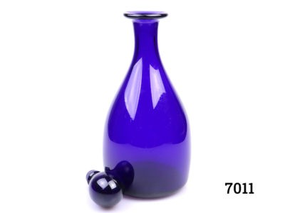 Vintage Thomas Webb Bristol blue decanter. Small chip on the collar. Measures 105mm in diameter at base and 50mm in diameter across the top. Photo of decanter with stopper removed seen from an eye level angle with stopper placed to the bottom left of decanter base