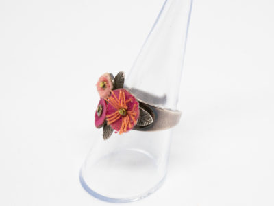 Pink flowers costume jewellery ring. Sweet white metal adjustable ring with a trio of different pink coloured felt flowers. Made in France by Taratata. Unadjusted size P / 7.5 Ring front measures approximately 20mm by 15mm.  Photo of ring on a clear display stand and seen from a side angle