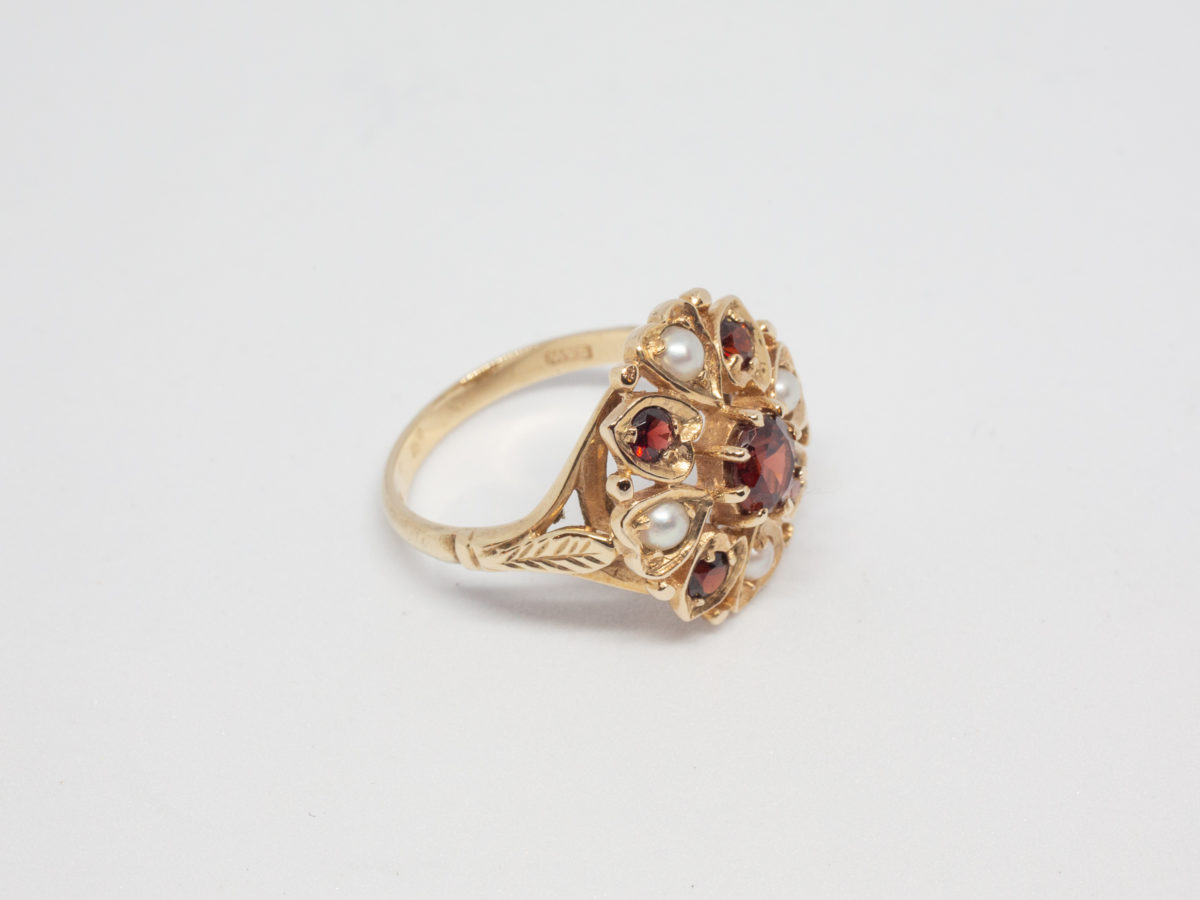 Vintage 9 karat gold ring with garnets and seed pearls. c1959 London assayed 9 karat gold ring with round cut garnet to the centre surrounded by 4 small garnets and seed pearls set on heart shaped petals with leaves on shoulders. Ring size O.5 / 7.75 Ring front measures approximately 18mm by 16mm. Photo of ring on a flat surface with ring front facing right & showing the leaf on shoulder