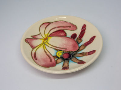 Moorcroft Frangipani Plumeria dish. Small round dish in cream with frangipani plumeria blossom. Stamped Moorcroft to the back and dated c1999. Measures 120mm in diameter and 75mm at base. Photo of dish with frangipani image shown from a side angle