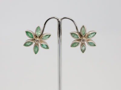 Modern sterling silver, emeralds and sapphire stud earrings. Very pretty pair or sterling silver stud earrings in a floral design with oval cut emeralds as the flower petals and a single round cut white sapphire to the centre. Earring front measures approximately 20mm in diameter. Main photo of both earrings displayed on a stand and shown from the front straight on.