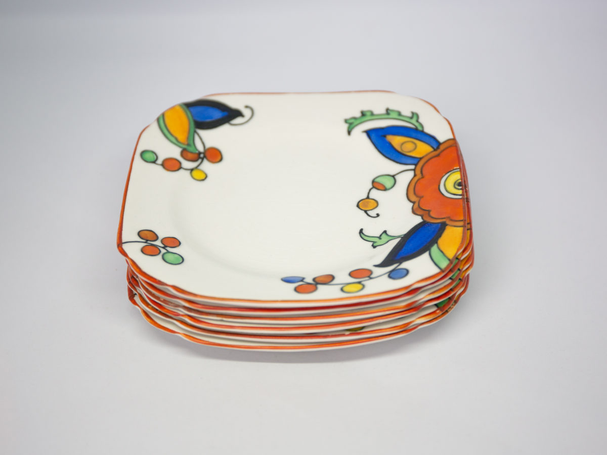 Art Deco sandwich set by Charlotte Rhead. A pretty and practical set comprising of a serving plate/tray with 2 shallow dish shaped plates connected with a handle to the centre and 6 small square side plates. Hand-painted in cheerful bright vibrant colours to cheer up any occasion. Design number 4133 'New Jazz' pattern c1928-1929. Each piece marked with the green Burleigh ware stamp with Rhead signature on the base of the serving plate. Visible crazing to each piece and some wear around the edge, especially around serving plate. Each small plate measure 145mm square and the serving plate/tray measures 330mm long,150mm wide and approximately 50mm tall by handle. Photo of the 6 small side stacked on top of each other plates