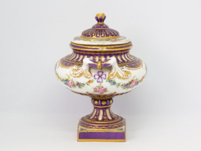 Antique Sèvres 2 handled urn with lid. Beautifully handmade and hand-painted Sèvres urn with 2 handles and lid in royal purple and gilt with floral design throughout. Some gilt wear mostly on the rim of the lid otherwise in excellent condition. Late 19th century. Base measures 55mm square, width at widest across handles 130mm, opening at top 72mm in diameter. Photo of urn from an eye level angle and shown from side angle with one of the handles in the centre foreground.