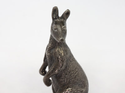 Antique Stewart Dawson kangaroo ornament. A small heavy kangaroo in standing position by Stewart Dawson. Assumed solid silver plate although no visible hallmarks whatsoever. Signed Stewart Dawson to the base of tail. c1900s Measures 85mm at longest from tip of foot to tail, 28mm at widest across hips and 98mm at tallest from ground to tip of ear. Close up photo of the top half of kangaroo with face turned straight to camera.