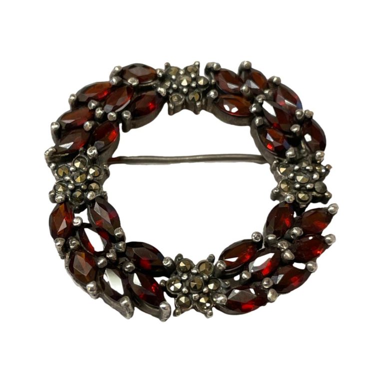 Main photo of silver garnet circular brooch shown from the front. The pin is visible going across horizontally in the background. The brooch is a wreath like shape and beautifully put together with a festive feel about it.