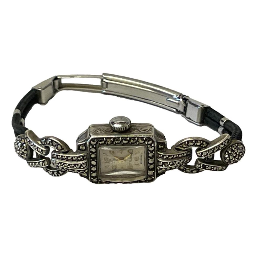 Watch laid on a flat surface shown with face in the foreground and clasp at back shown closed. The side areas are black leather.