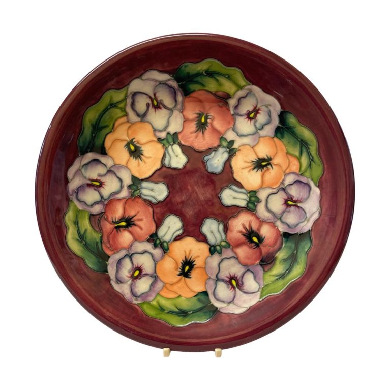 Main photo of the plate shown displayed on a small plate stand and forward facing. The base colour is a maroon with the pansies in a wreath like formation in lilac, orange and pale red with green leaves. Very striking colour palette.
