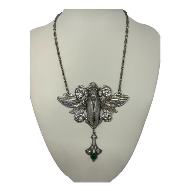 Silver plate cicada necklace by Lelong Designs, London. Inspired by the Art Nouveau era, this statement necklace has a large pendant which is attached to the chain. It houses a cicada nestled to the centre of tree seeds with an extension to the base. A single green onyx cabochon adding to the Art Nouveau style. The pendant area measures 90mm at widest with drop length of 100mm. Main photo of necklace displayed on a stand and seen with pendant forward facing.