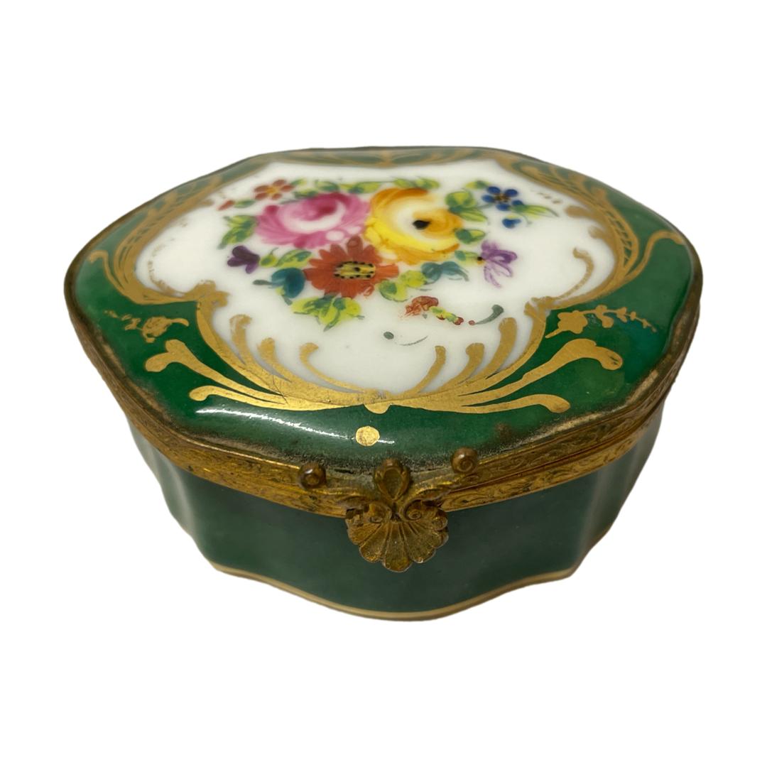 Small porcelain box by Paris. Lovely small forest green scallop shaped box by Paris porcelain. Hand-painted with colourful flowers framed within gold to the top with a single pink rose to inside of the box and lid. Finished in a brass edge with small scallop opener to the front. Signed to the base. Some minor signs of wear to the brass. Main photo of box seen with the opening front in the foreground and shown from a slightly raised angle to show the hand-painted floral top of the box.