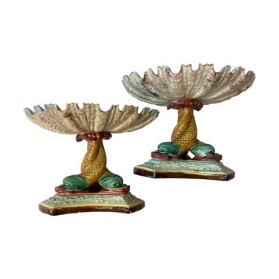 Pair of Art Nouveau sweetmeat dishes. Colourful pair of late Victorian Art Nouveau majolica sweetmeat dishes. Each triple footed dish has 3 dolphins entwined to create the column which houses a scallop shaped shell like dish. The colour combination of turquoises and sea greens against pinks and yellow give these pieces are true under the sea look. Some chips to the paint and some scallop edges. Each dish measures approximately 105mm by 105mm at the base, 103mm tall and 143mm in diameter to the dish at top. Main photo showing both dishes from an eye level with focus on the entwined dolphin columns and detail to the underside of the scallop shell. One dish is slightly to the foreground and they are placed diagonally side by side.