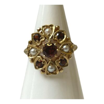 Garnet and pearl ring on 9 karat gold. Vintage ring with a round cut garnet to the centre and framed with alternating pearl and garnets each in a heart frame setting. Has a Tudor or Queen of Hearts look about it. Fully hallmarked for London assay and made by Slade and Wolf c1959. Ring size O / 7.25. Ring weight 4.4grams. Main photo of ring displayed on a cone shaped stand and facing with front forward. The largest garnet is to the centre with the small pearls North, South, East and West and the smaller garnets in between.