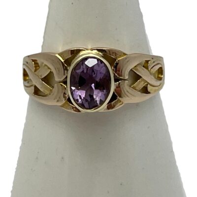 15 karat gold ring with amethyst. Vintage ring with a deep lilac oval cut amethyst set in 15 karat gold. Celtic knotwork like shoulders add an extra special touch to this pretty ring. Hallmarked 15ct to inside band. Ring size M.5 / 6.5. Main photo of ring displayed on a cone shaped stand with ring front facing forward.