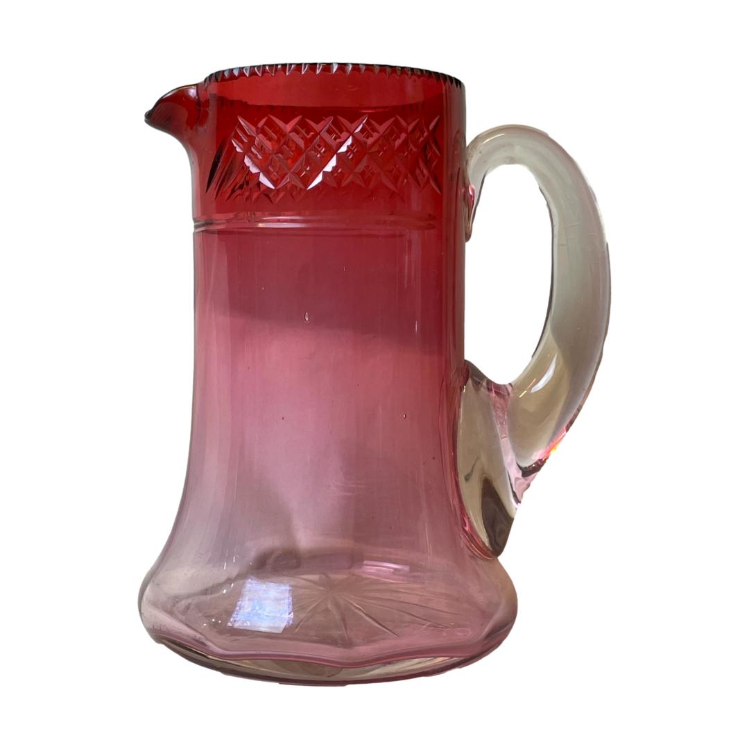 Vintage cranberry glass jug. Handblown mid sized etched cranberry glass jug. Etched star burst pattern to the almost clear base and criss-cross design to the top. Measures 105mm in diameter at base and 130mm at widest from tip of spout to handle. Main photo of jug seen from a near eye level with the spout to the left and handle to the right. The cranberry colouring of the jug is darkest at the top and graduating downward to clear.