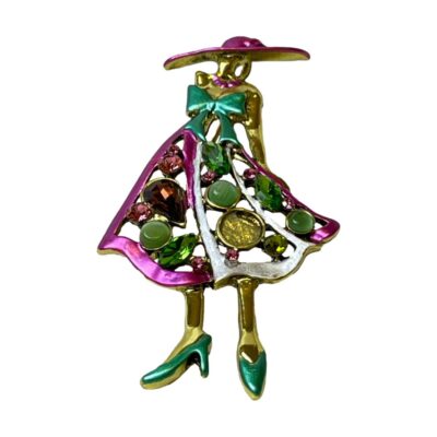 Modern brooch/pendant of a vogue lady. Fun brooch of a fashionable lady dressed in a vogue style. Main colours of attire in turquoise green and magenta pink strikingly set against a gilt metal body. The skirt of the dress is made up of different shaped glass stones which have colours complimenting the 3 main colours of the brooch. Photo of brooch taken from a lower angle looking upward to focus on the skirt and its multiple stone decorations.