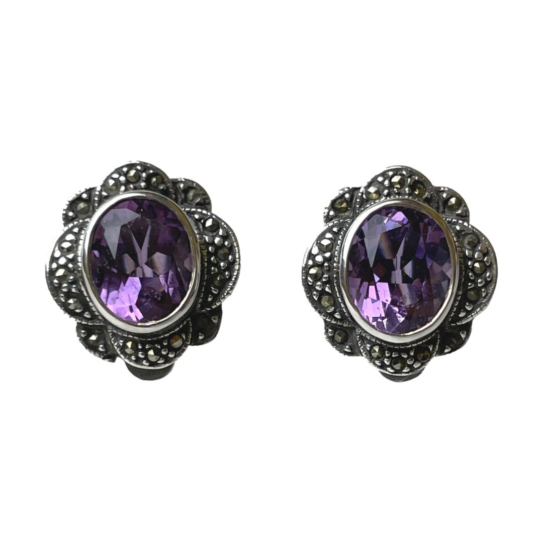 Vintage clip-on earrings with amethyst. Sterling silver clip-on earrings with a good sized oval cut amethyst stone to the centre with marcasite surround. Hallmarked 925 for sterling silver to the back. Both clips in good working order. Earrings measure 20mm long by 16mm wide. Amethyst measures 10mm by 8mm. Main photo of earrings seen with the front forward facing. The oval amethysts are vertical with the marcasite frame arranged in a wavy flower like pattern around it.