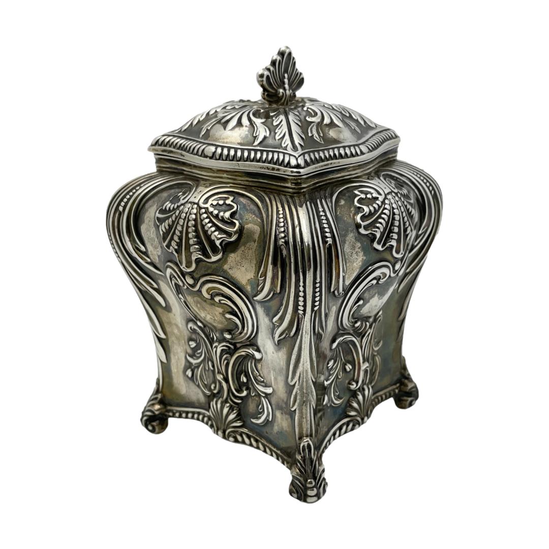 Main photo of caddy seen with one corner to centre foreground displaying 2 sides of the square caddy. The incredible decorative detail of the silver work is shown with scallops and swirls amongst others.