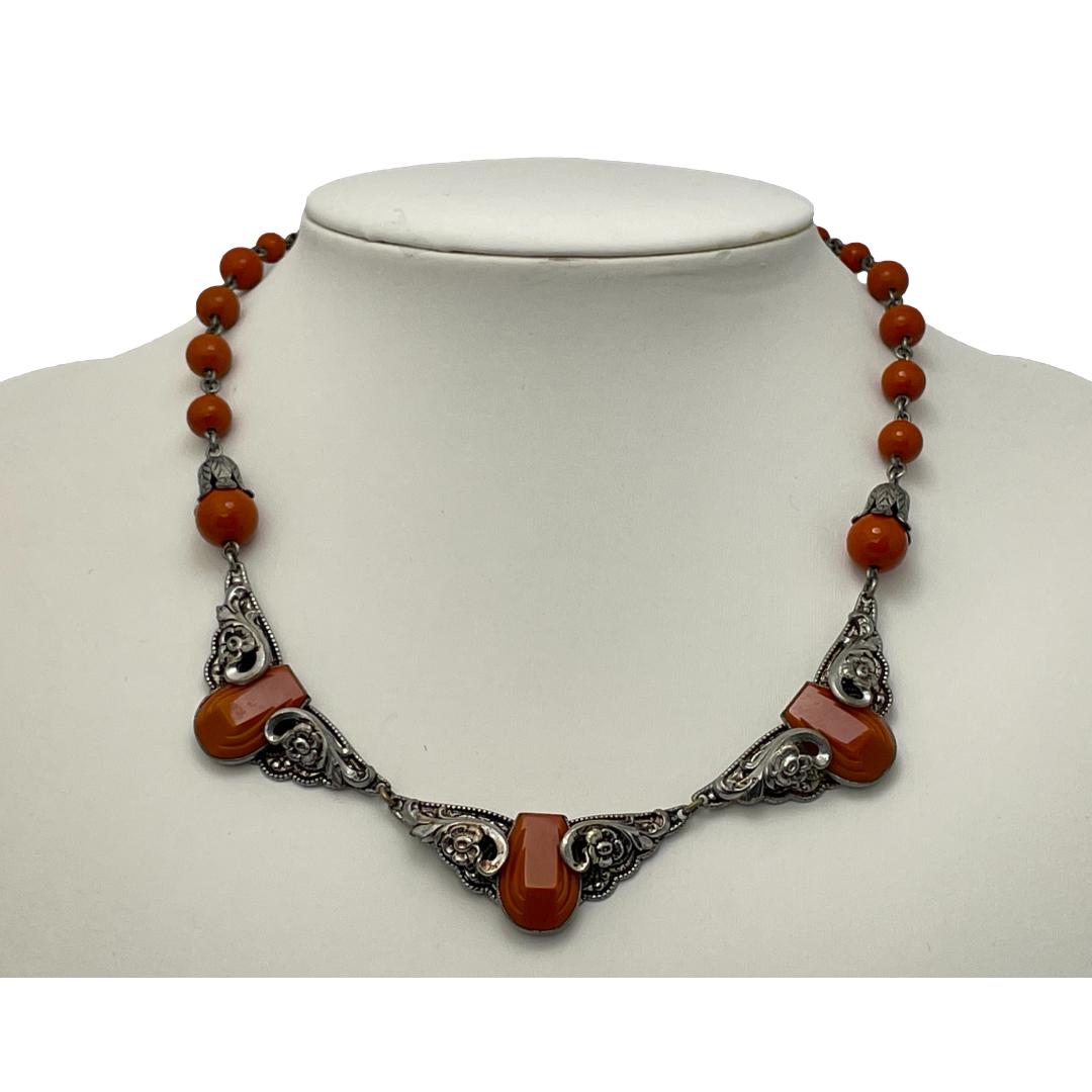 Main photo of necklace displayed on a stand and seen from the front. There are 3 wide bunting like panels at the front which are of a floral theme with a piece of carved red jasper to the centre of each.