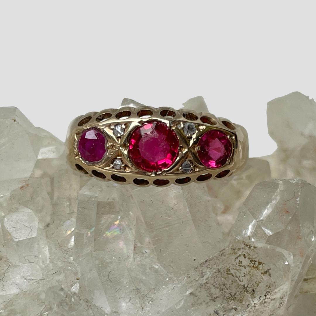 Main photo of ring displayed on top of a cluster of quartz crystal. The ring front is forward facing. The largest of the 3 rubies is to the centre with the smaller ones to either side. Each ruby is separated with 2 small diamonds chips one above the other. The rubies are a deep pink colour.