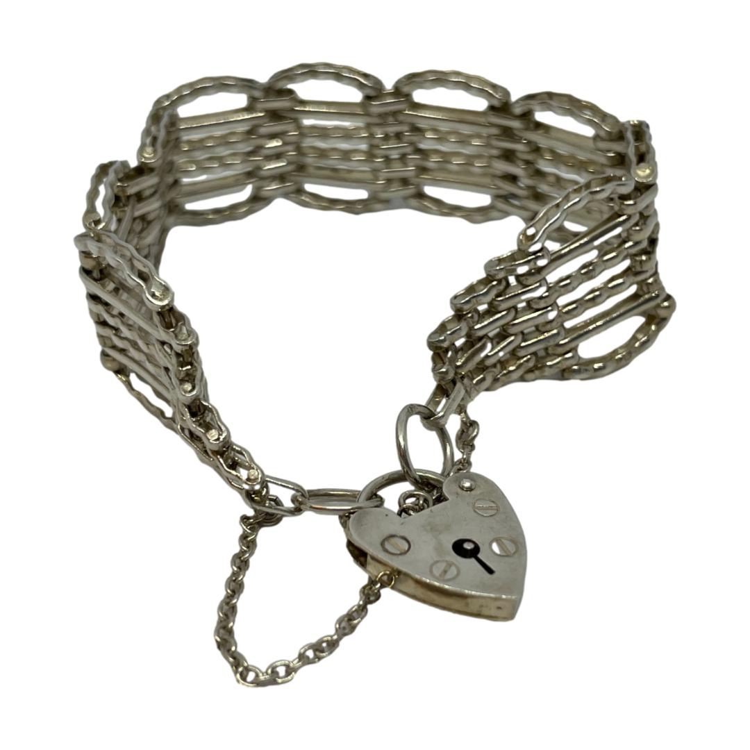 Main photo of vintage silver gate bracelet displayed in a circle on a flat surface with the heart clasp in the centre foreground. The main body of the bracelet is shown balanced upright with the panels positioned to keep it upright. The clasp is closed. There are 10 gate panels to this bracelet and each panel has 6 bars; 4 to the centre and a curved one above and below the central 4. Each panel is then separated with 5 links making a very secure setting. The end of each of the panels comes to a point with safety chain attached to ring hooks which the heart clasp threads through. Quite an unusual gate bracelet in its design.