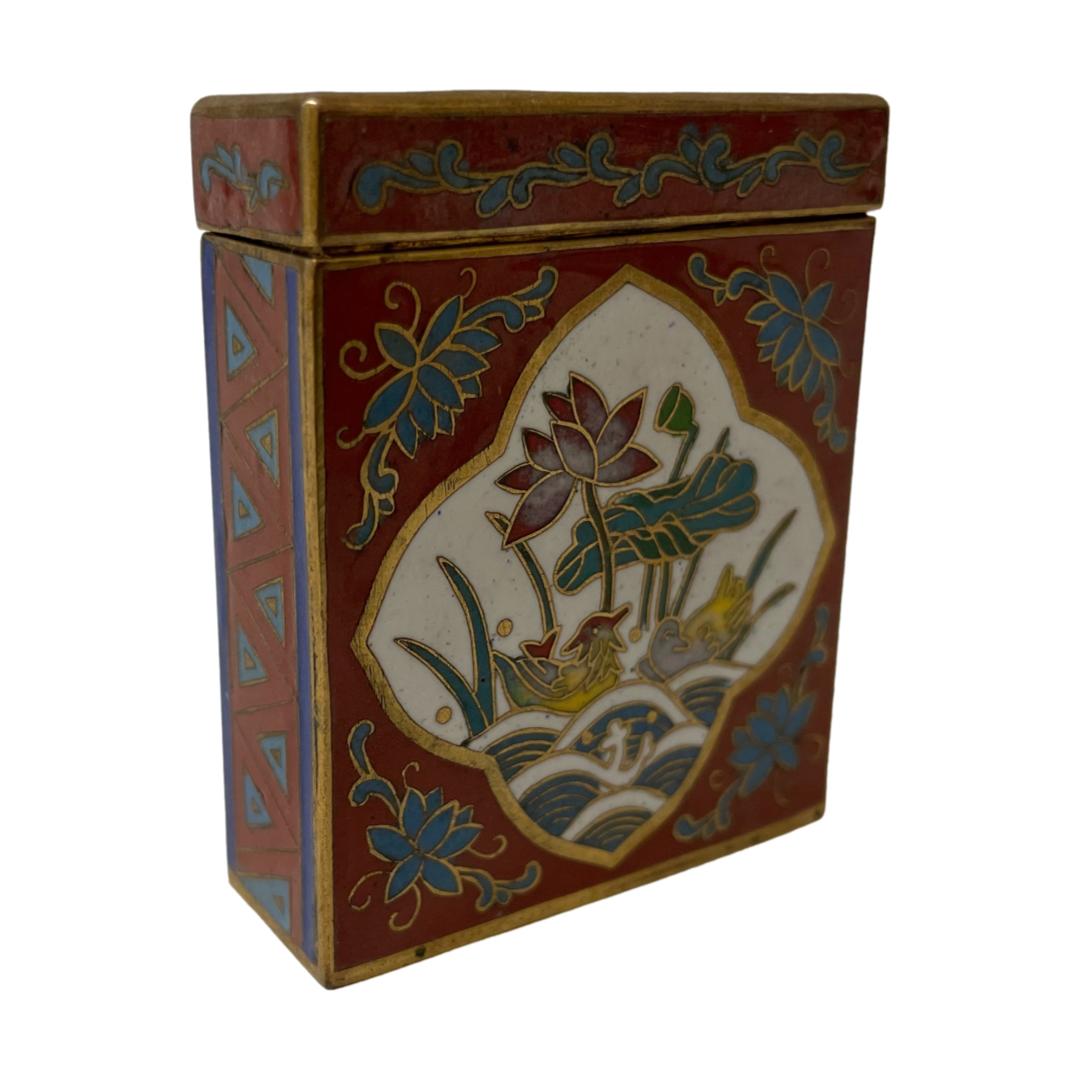 Main photo of small vintage cloisonné box displayed at an angle so one wide decorative side is facing towards the bottom right corner of photo and a thinner side facing bottom left corner to show the depth. The image on both decorative sides are the same set in a wide diamond shape to the centre are a pair of courting Mandarin ducks below a blooming lotus flower. The main colour of the box is a deep red with floral design around the image in air force blue and small triangles along the thinner side and to the lid. The lid is lift off and not hinged.