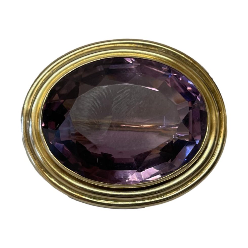 Main photo of 14kt gold amethyst brooch displayed against a white background and in close up. The large oval amethyst is a rich purple shade and is neatly faceted around the edge. The gold around the stone has a classical look with double beading. The colours marry well together.