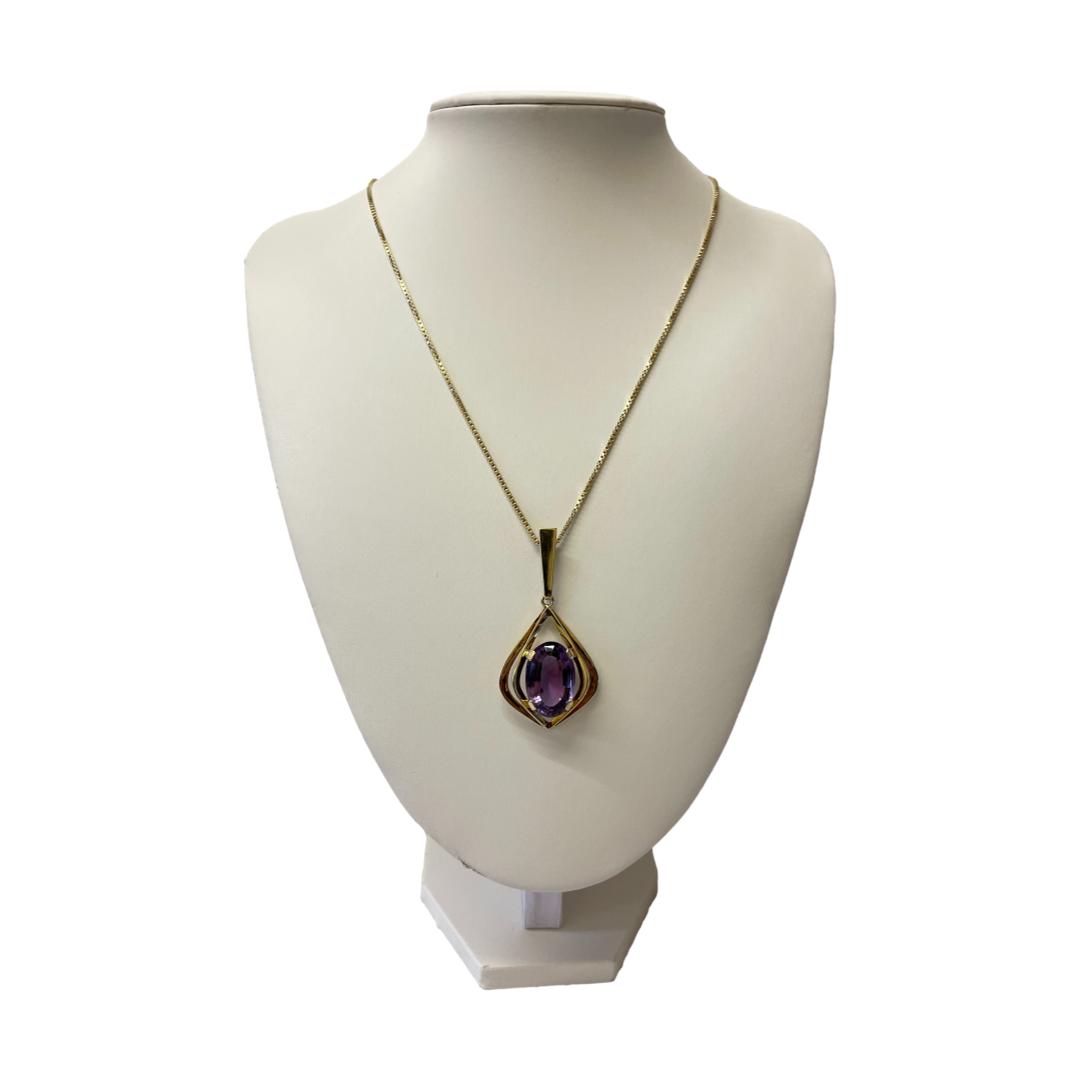 Main photo of 14kt chain & amethyst pendant displayed on a necklace display stand of an off white colour and shown with the front of necklace facing forward. The pendant has a fairly long bail shaped like the top of an exclamation mark and the main shape of the main pendant is a little like a teardrop with a pointed tip and the end. There are 2 'frames' of gold around the large oval cut amethyst, the inner frame has 4 clips which hold the amethyst in place and which are positioned approximately at 2, 5, 7 and 10 o'clock. A second frame identical to inner frame just wider and longer, sits outside the inner frame. The chain is a nice box chain with good thickness which compliments the pendant perfectly.