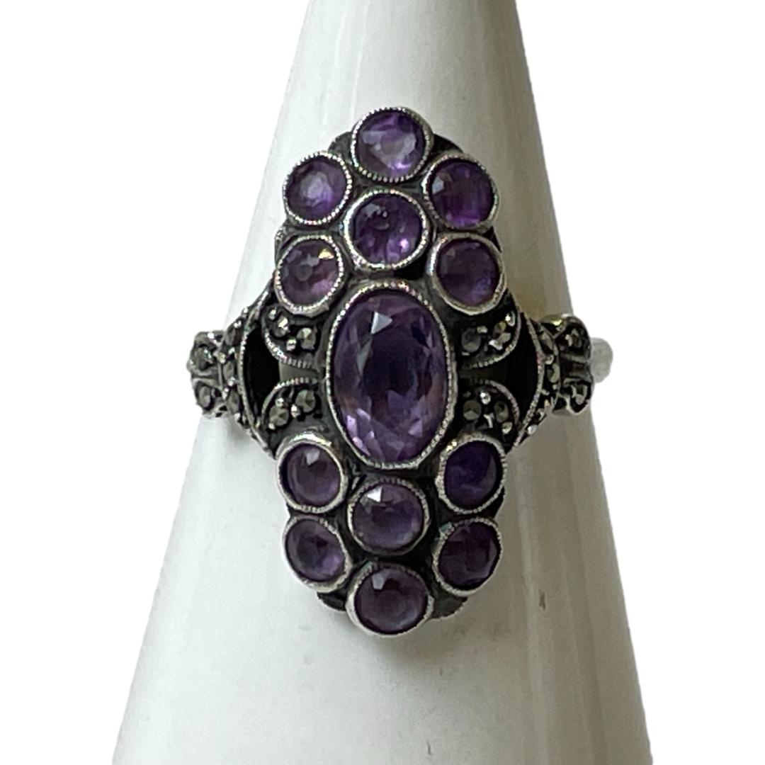 Main photo of 935 silver amethyst ring shown displayed on a cone shaped stand and with the ring front forward facing. The oval central amethyst sits vertically with the 6 smaller round cut amethysts placed 2 up in a row of 3 at the top and below the central stone making this a vertically long ring. To the left and right of the central oval stone is a little marcasite then a small gap followed by the shoulders which also have a little marcasite. The band is plain silver. The silver looks fairly dark in this photo and could probably do with a little more cleaning!