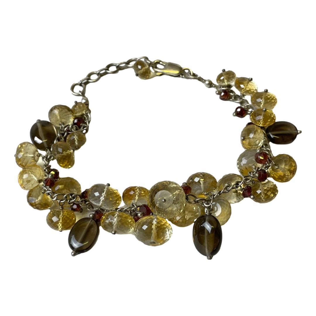 Main photo of sterling quartz crystal bracelet displayed on a flat surface in a circle with the clasp closed. The quartz crystal pieces along with the small cranberry coloured beads are faceted while the smoky quartz beads are smooth ovals. It has a nice neutral earthen colour scheme.