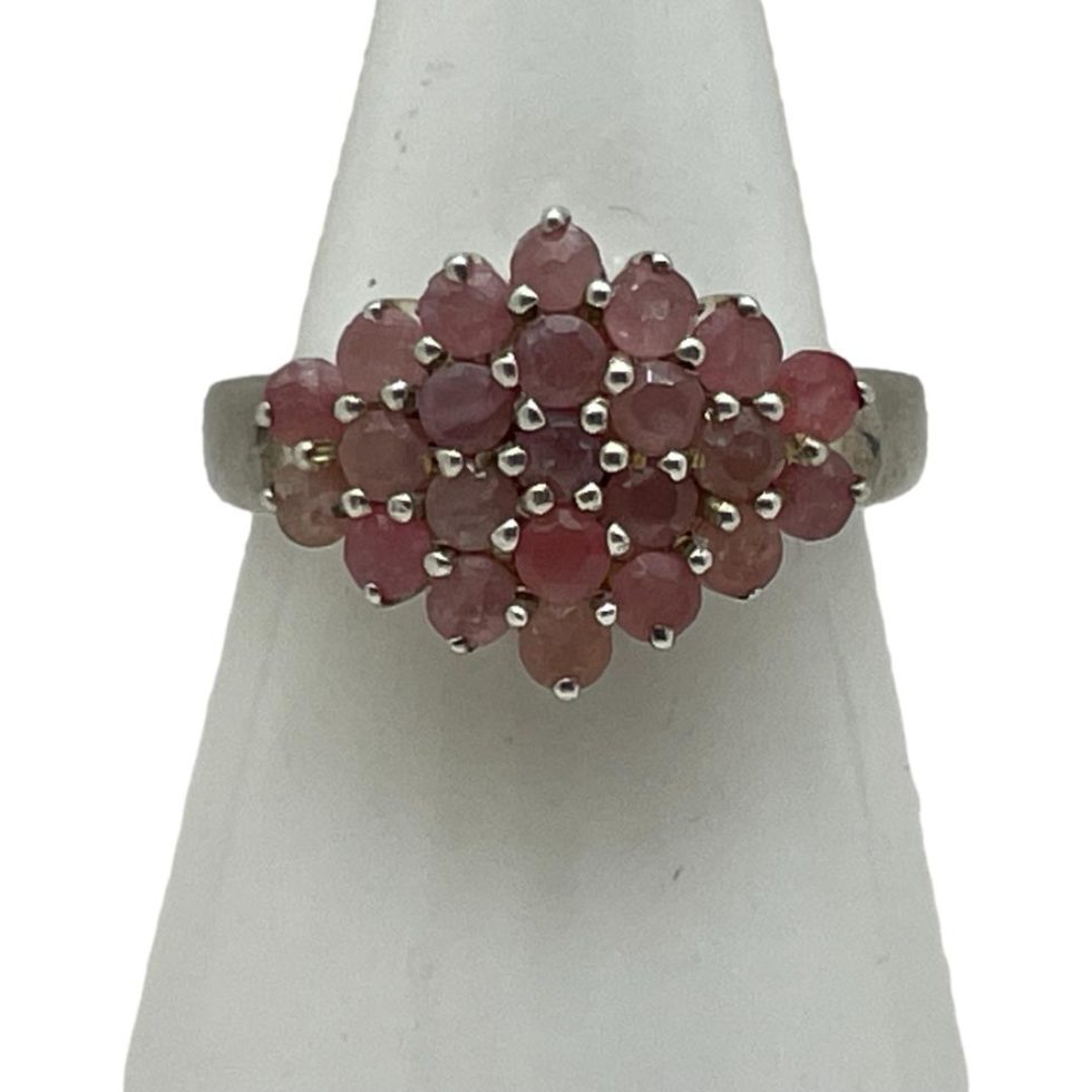 Main photo of silver &ruby cluster ring displayed on a white cone shaped stand and seen with the ring front forward facing. This is quite an unusual ring in that the cluster is side wide rather than circular. The rubies are a sweet pale pink and red colour mix which marries well with the silver.