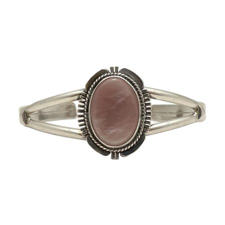 Main photo of Navajo signed cuff bracelet displayed on a flat surface against a white background with the front of bracelet at centre and forward facing. The cuff has a split band at front with an oval central frame which houses the dusky pink coloured natural shell. The frame is a very recognisable Navajo design with 'feathered' edges at the 4 compass points and intricate rope twist around the stone.