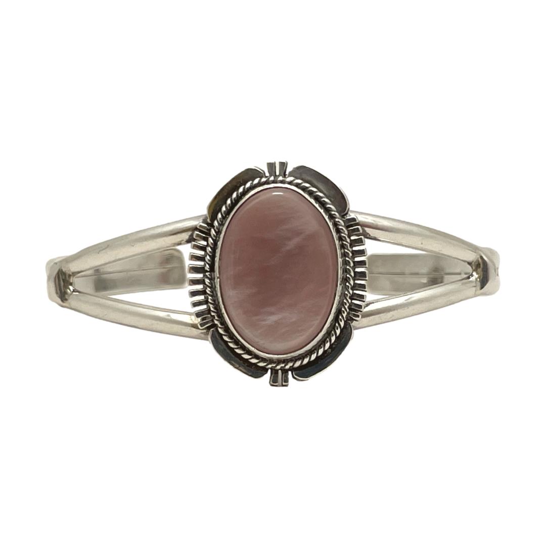 Main photo of Navajo signed cuff bracelet displayed on a flat surface against a white background with the front of bracelet at centre and forward facing. The cuff has a split band at front with an oval central frame which houses the dusky pink coloured natural shell. The frame is a very recognisable Navajo design with 'feathered' edges at the 4 compass points and intricate rope twist around the stone.