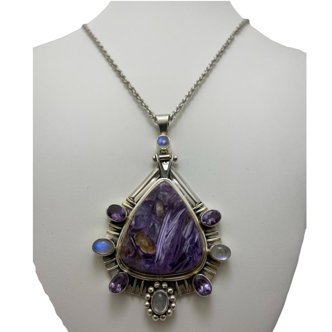 Main photo of statement charoite pendant necklace displayed on an off white coloured stand and displayed with pendant forward facing and stealing the show! Charoite is a lavender purple colour with a pearly lustre and unusual swirling fibrous appearance. It is found only in Siberia. This charoite stone has a triangular shape set in silver. Around the wider end of the triangle, there are 7 silver protrusions; top 2 with amethyst, then rainbow moonstone, then back to amethyst and finally at the bottom encased in a floral silver setting, another rainbow moonstone. At the bail, there is another rainbow moonstone to settle the balance. The chain is a good length and thickness but frankly pales in comparison to the gorgeous pendant.