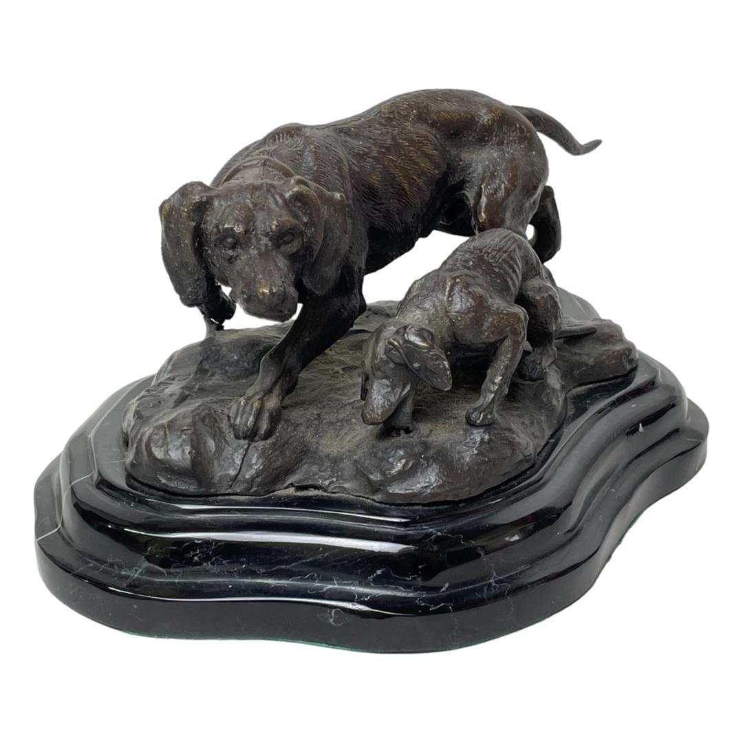 Main photo of signed bronze hunting dogs seen with the adult dog in the background facing camera and appears to be guarding the pup in the foreground while it drinks. The bronze is set on a heavy slab of black marble.