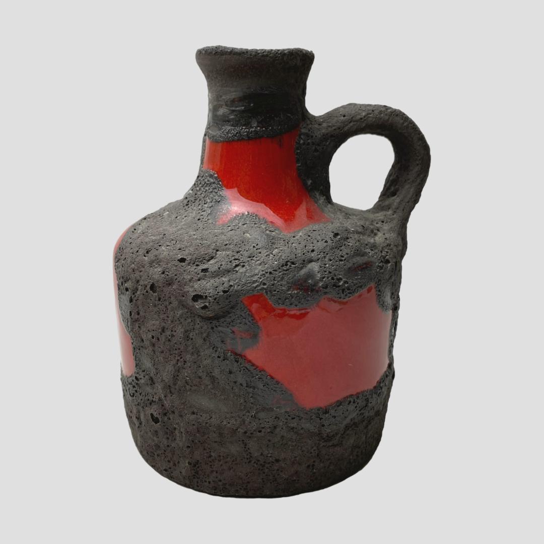 Main photo of Marei Keramik fat lava jug seen from an eye level with the handle to the right of shot. The main body is a vibrant glossy red with black lava laid over the top. This is a very striking rustic piece.