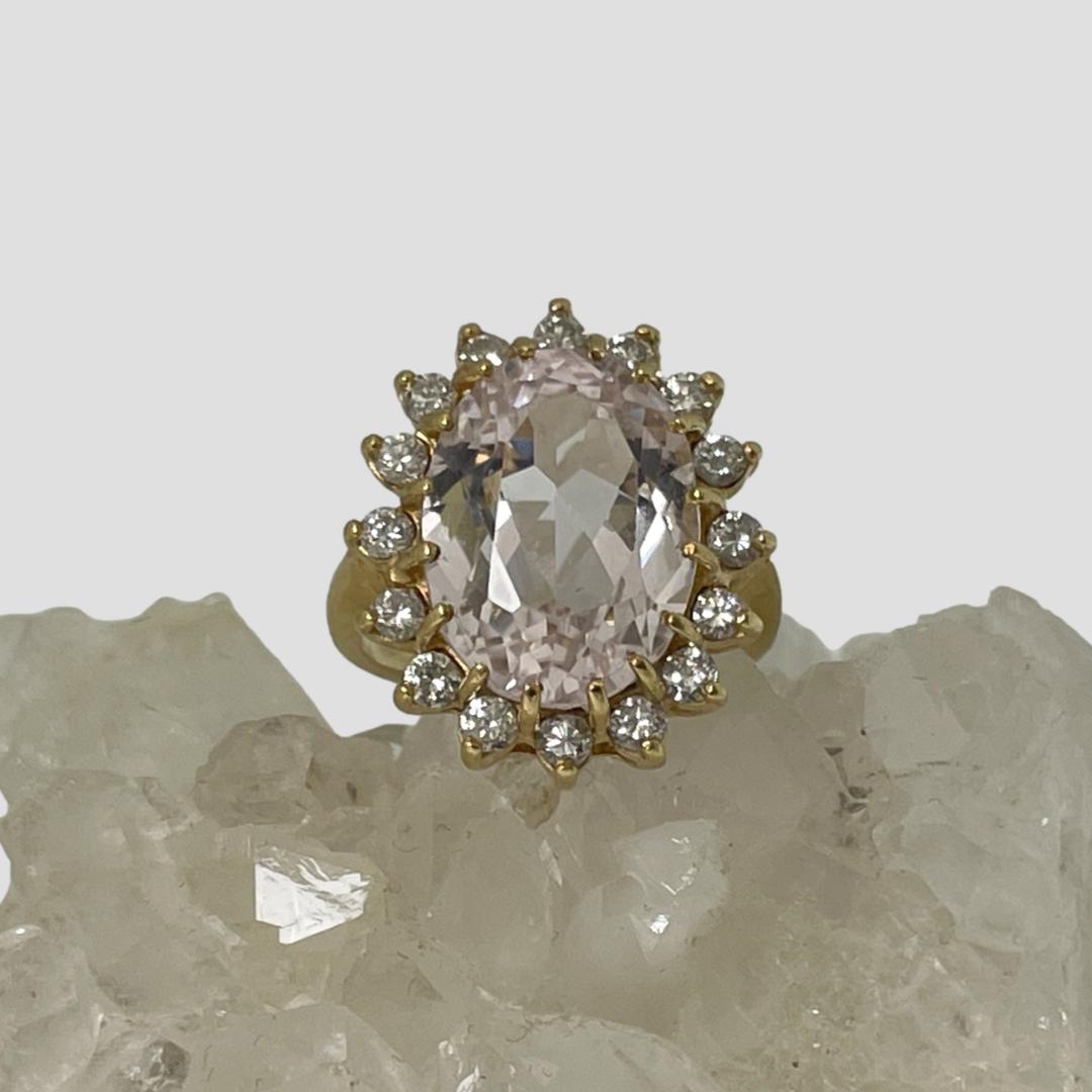 Main photo of rose quartz diamond ring displayed on top of a rock of natural quartz crystal with the ring front forward facing. An unusual pairing, the large oval rose quartz is beautifully faceted with the 16 diamonds set all around on an external setting.