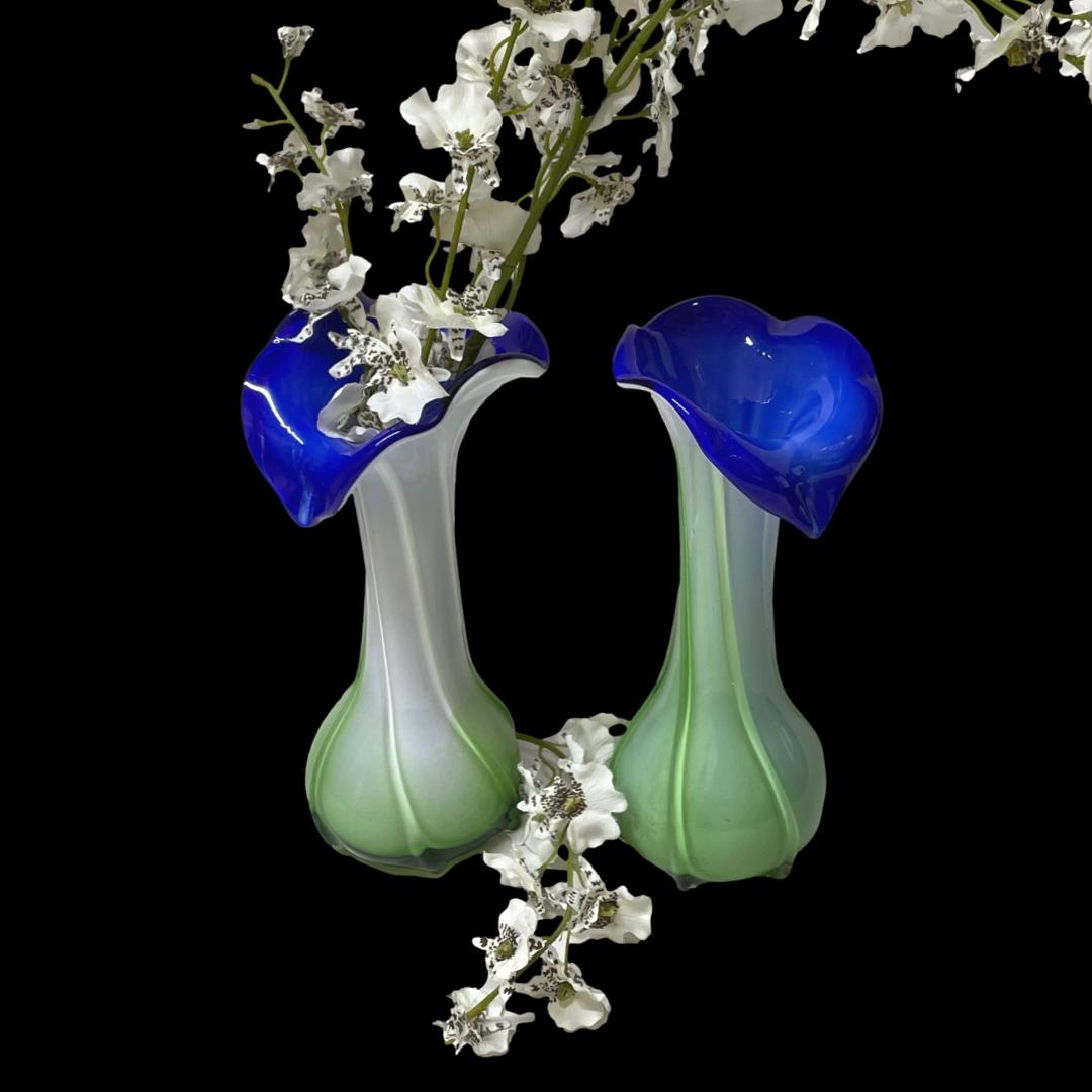 Main photo of the 2 calla lily glass vases displayed side by side and from a slightly raised angle looking diagonally down. The main body of the vases is a mix of white and pale green set in a light swirl. The mouth of the vase opens out and protrudes hanging outward with interior a beautiful vibrant blue. The vases on the left has a spray of white orchids in with another spray laid in between the 2. Against a black background, these vases are more striking!