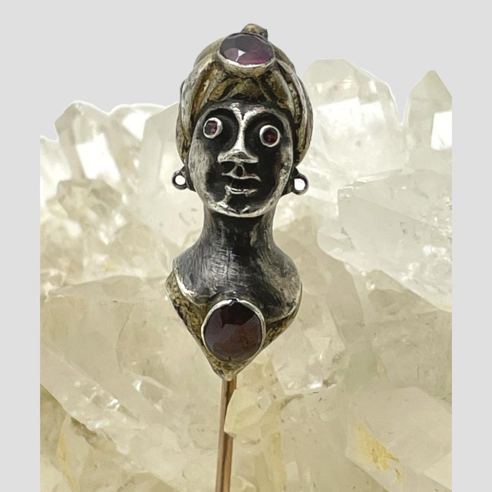 Main photo of Austro-Hungarian tie/stick pin showing the top part of the pin which has the bust of the lady. She has a tight fitting turban like hat on with an amethyst to the centre and amethyst eyes. She is also wearing a necklace with a large carnelian cabochon. The turban has gilt and silver stripes and she is wearing a gilt gown although only the neckline area can be seen. She has a strange look about her face like she is a little crazed or drunk - a very quirky piece indeed!