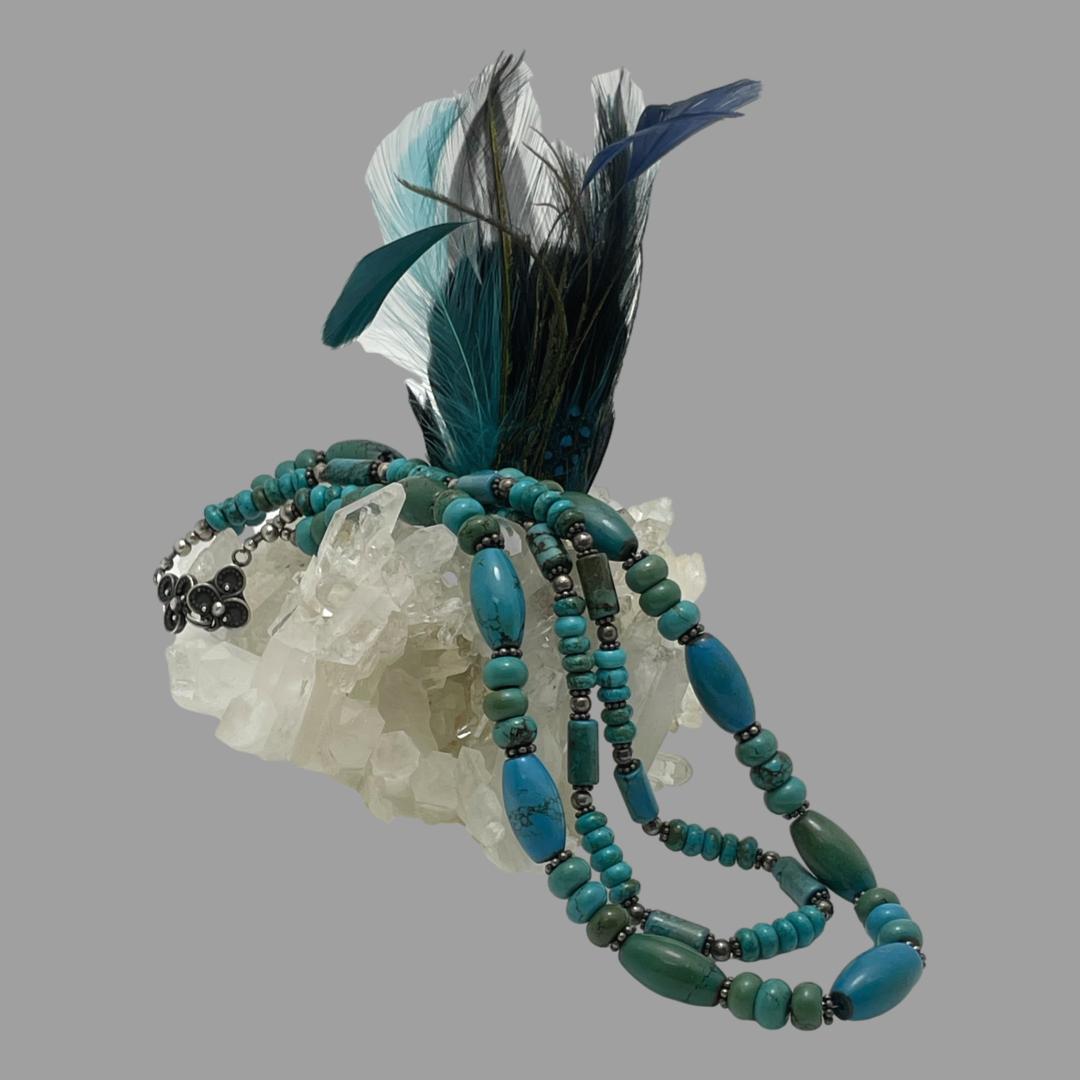 Main photo of 2 strand turquoise necklace displayed with the clasp end on top of a rock of natural quartz crystal and other end draped down to the right. A cluster of blue feathers are seen behind the necklace. The longer strand of the necklace has larger oval turquoise beads each separated with 3 flat round beads. The shorter of the 2 strings has smaller cylindrical tube beads with 5 small flat round bead separators. The colour range of these turquoise beads is incredible from a darker turquoise to green and of course regular turquoise!