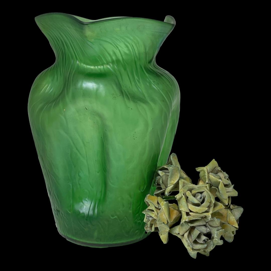 Main photo of Johann Lötz Witwe vase seen from an eye level against a black background. The green colour of this vase is breathtakingly beautiful with a light iridescence. Interesting pentagonal shape with dips and markings. A small posy of blue green roses are laid to the right of the vase.