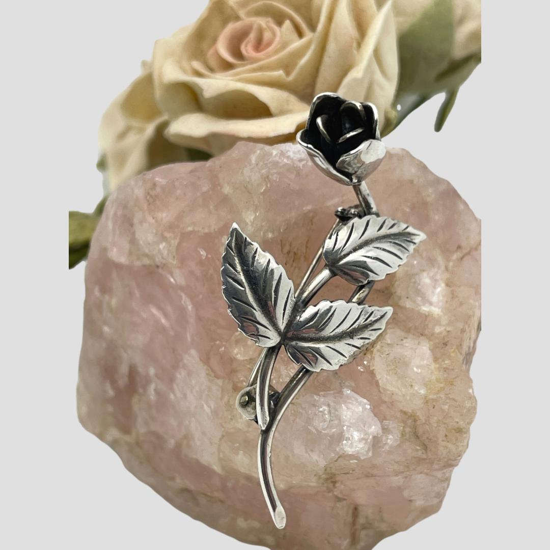 Main photo of SCF Denmark rose brooch displayed on top of a small rock of rose quartz with a pale pink rose in the background. This beautifully crafted brooch is purely of sterling silver with no other colours or materials involved. The brooch is a single rose stem which has a curved structure. From near the base of the stem a smaller stem comes out with 3 leaves attached. Behind this stem of leaves is the brooch pin cleverly concealed! A second curve up the stem ends with a single rose open but not fully.