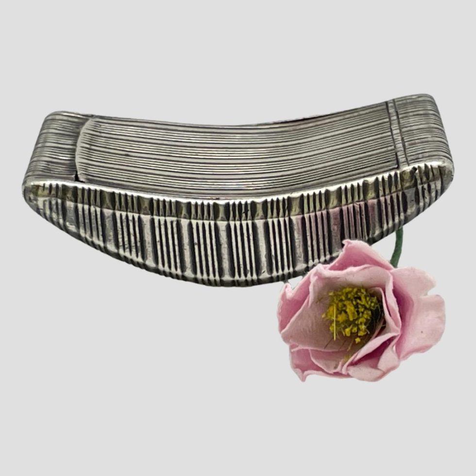 Main photo of George III snuff box seen from an almost eye level. This snuff box has a curved bottom for ease of fitting into pockets - it could also be used as an executive toy on the desk rocking back and forth. This photo shows the box from one of the sides on display showing the engine turned decoration which extends across the whole box. The lid is well camouflaged as it sits flush with the curved top but in this photo it is to the left with hinges visible to the right. A single pink flower rests to the right foreground of the box.