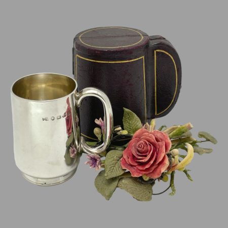 Main photo of antique cased christening mug displayed outside of its fitted case. The mug is in the foreground to the left with the handle to the right. The case for the mug is closed and seen slightly to the right behind the mug. There is a little display red rose cluster on the right. The angle of the photo is from a raised angle looking diagonally downward peering into the gilt interior of the mug. The hallmark is visible on the outside of the mug by the top of the handle.