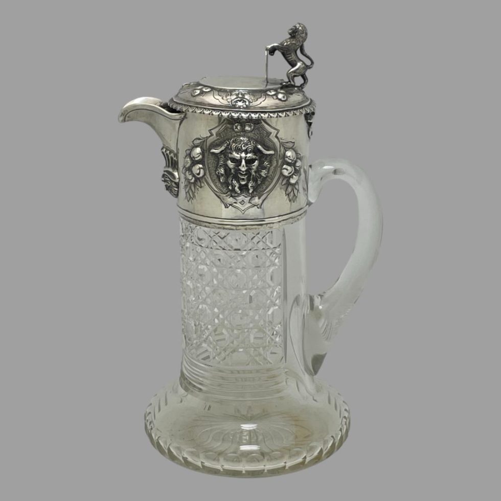 Main photo of Victorian crystal claret jug seen from an eye level with the spout facing to the left. The handle to the right is crystal and the top of it starts below the silver upper area of the jug. The lion is in profile and standing on its hind legs with its front paws resting on the shield with a cockerel on the front. The rest of the sterling silver top is decorated with berries and the face of Bacchus - the Roman God of wine. The crystal body of the jug is beautifully hob nail cut.