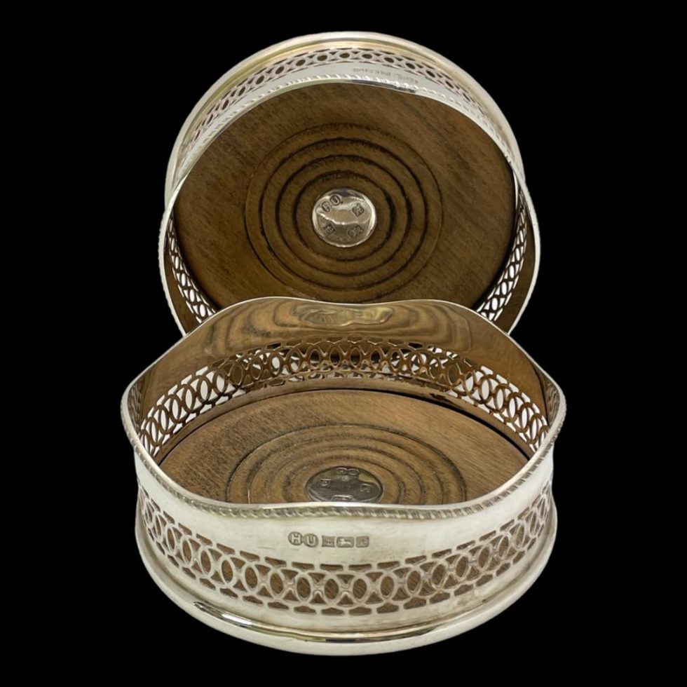 Main photo of vintage silver bottle coasters displayed with one laid flat in the foreground and other behind it upright showing the wooden centre. The coaster in the foreground has the hallmark visible at the front. These coasters are in good condition with very minimal signs of wear.