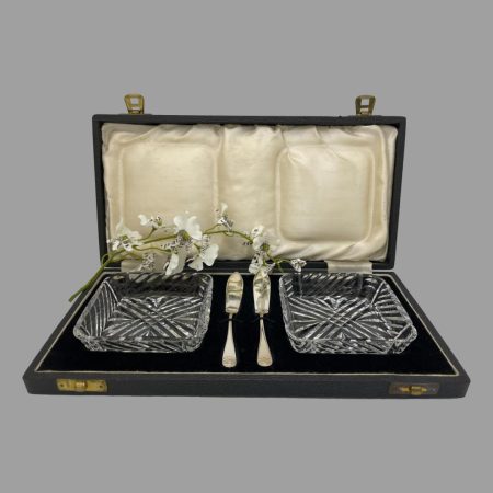 Main photo of cased butter dishes &knives displayed inside their original fitted case. The lead crystal butter dishes are to the left and right inside the case with the small silver butter knives slotted vertically at the centre. The case is in very good condition except for mild foxing of the silk to inside lid. A spray of white orchids is between the lid silk and fitted velvet area for display purposes only.