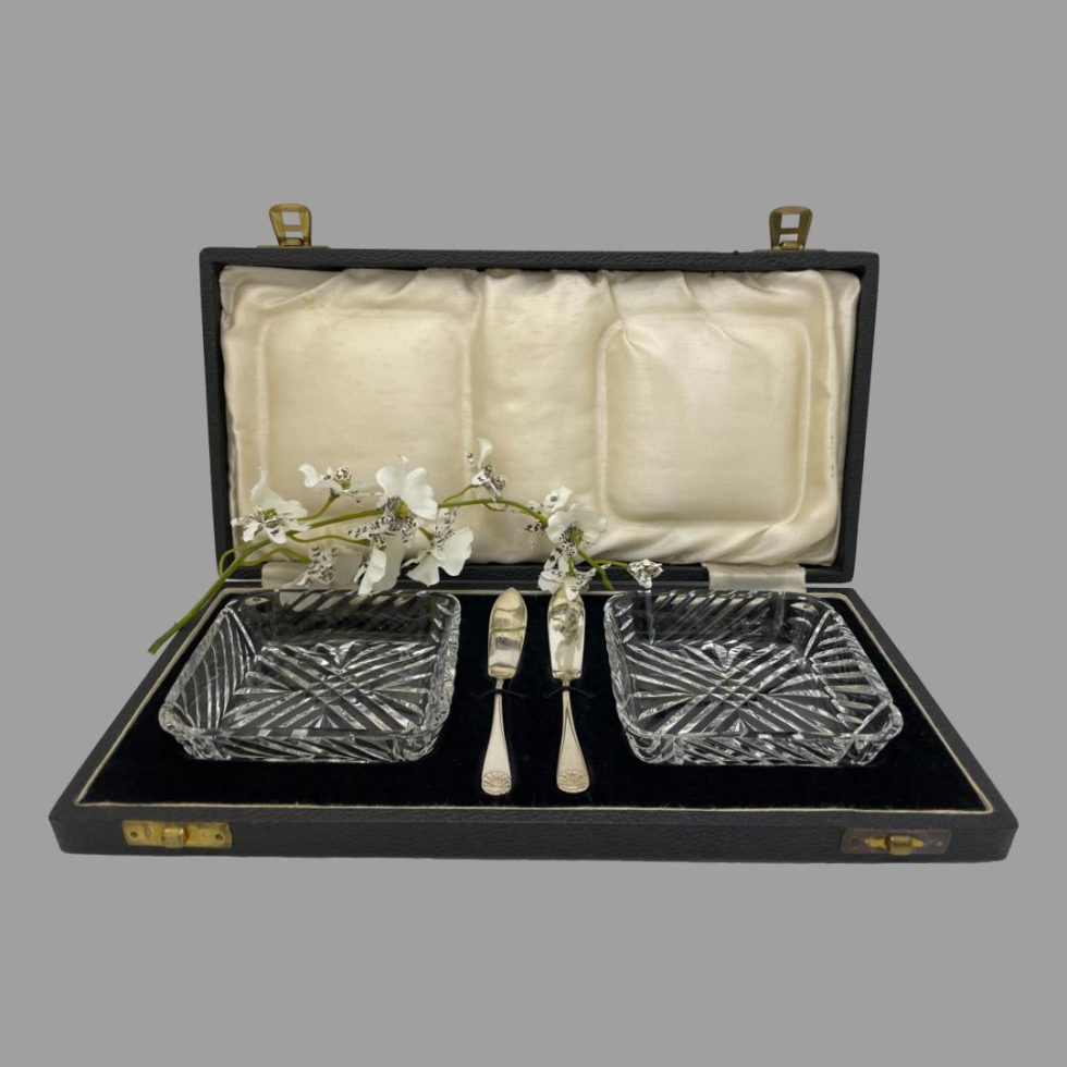 Main photo of cased butter dishes &knives displayed inside their original fitted case. The lead crystal butter dishes are to the left and right inside the case with the small silver butter knives slotted vertically at the centre. The case is in very good condition except for mild foxing of the silk to inside lid. A spray of white orchids is between the lid silk and fitted velvet area for display purposes only.