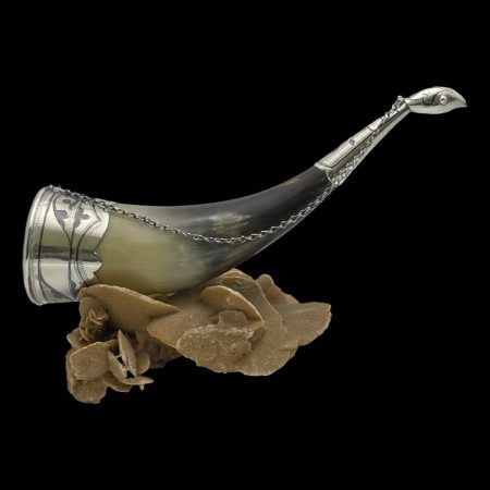 Photo of Russian horn drinking vessel displayed on top of a rock of natural desert sand rose. The pointed tip of the horn has a little silver covering which has a birds head at the end. The mouth end of the horn has a one and a half inch silver rim on the outside. A silver chains extends from the mouth of the vessel extending to the back of birds head at the tip making a handy carrying handle that can be hooked to a belt or hung up to dry after use. A perfect gift for the Viking in your life!
