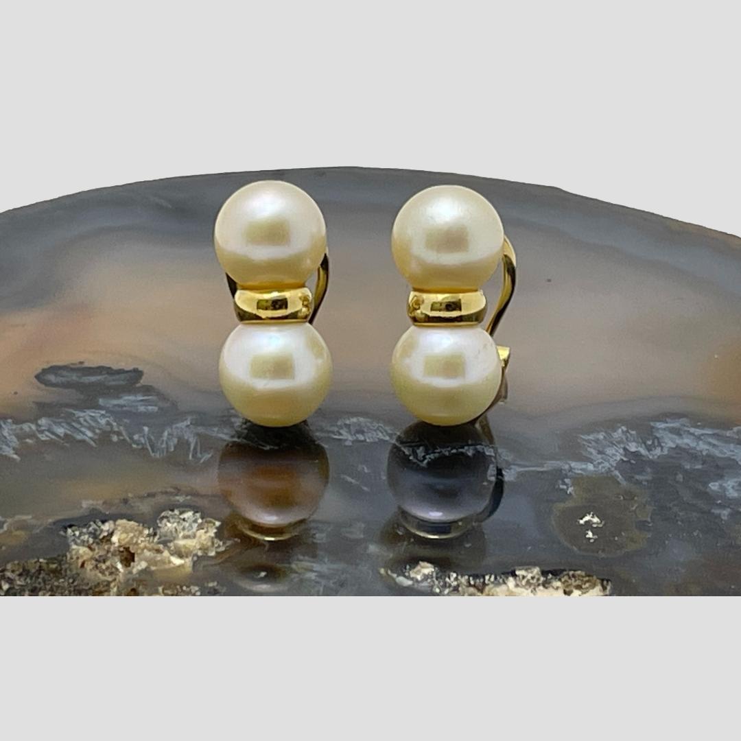 Main photo of gold and pearl clip-on earrings displayed on top of a slice of natural agate with the earring fronts forward facing. The pearls are set one above the other with a gold divider. A little like a figure of eight with an elongated mid section.