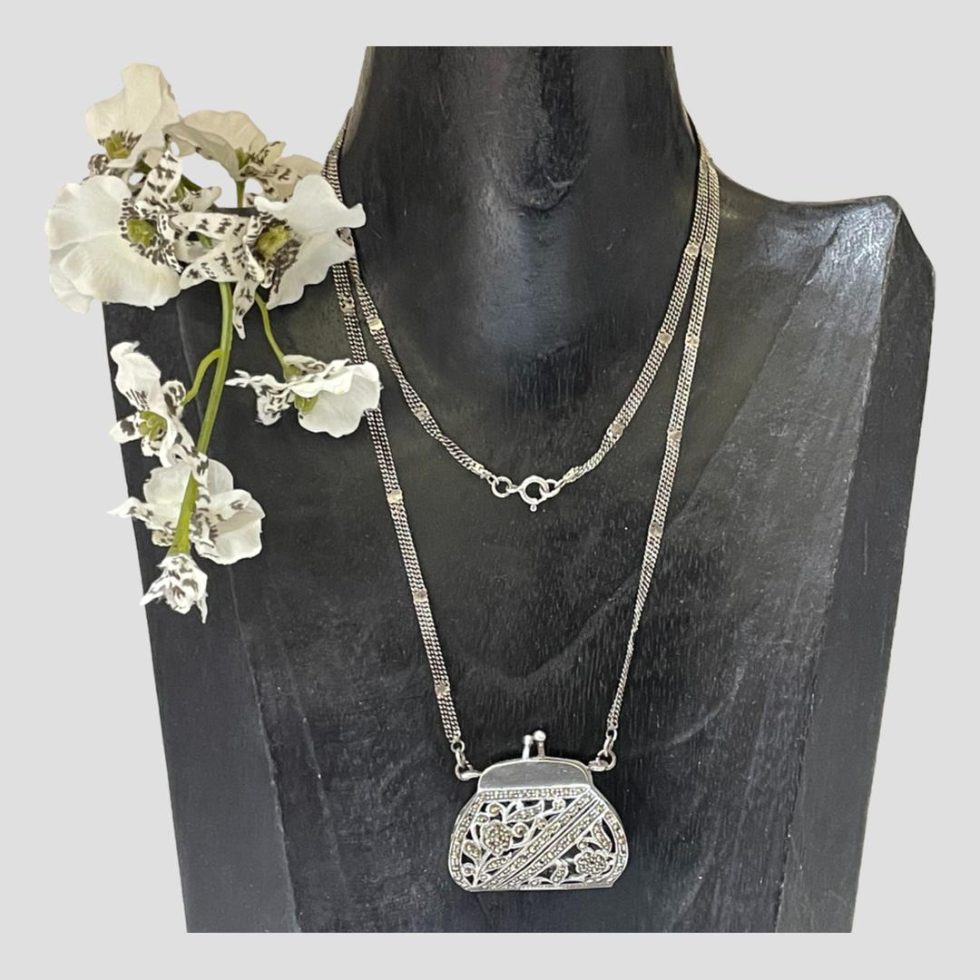 Main photo of silver purse pendant &chain displayed on a black wooden display stand. The chain has been doubled up as it is quite long. The purse is in marcasite of perforated design with a floral theme. The double chain is held together with silver clips so appears as one chain but is actually 2 clipped together. There is a spray of white orchids on the lefthand side shoulder of the display stand.