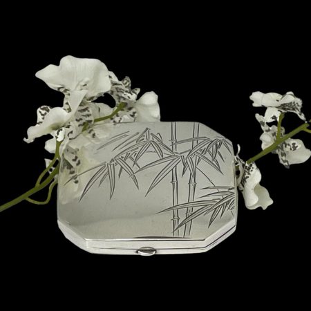 Main photo of Oriental 950 silver compact seen from a slight height looking diagonally down at the decoration on the lid cover. There are 2 bamboo stalks slightly on the right side of lid with the bamboo leaves over the top and one spray of leaves hanging over to the left hand side. The compact is set against a black background with a spray of white orchids placed around it for display purposes.